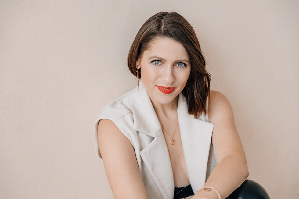 Portrait einer braunhaarigen Frau vor einer beigen Wand, sie trägt eine weiße Weste