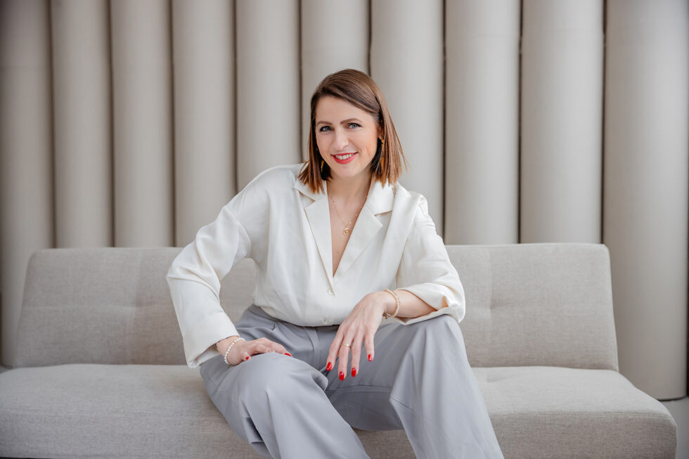 Businessportrait einer Frau mit weisser Bluse und grauer Jeans, die auf einem Sofa sitzt