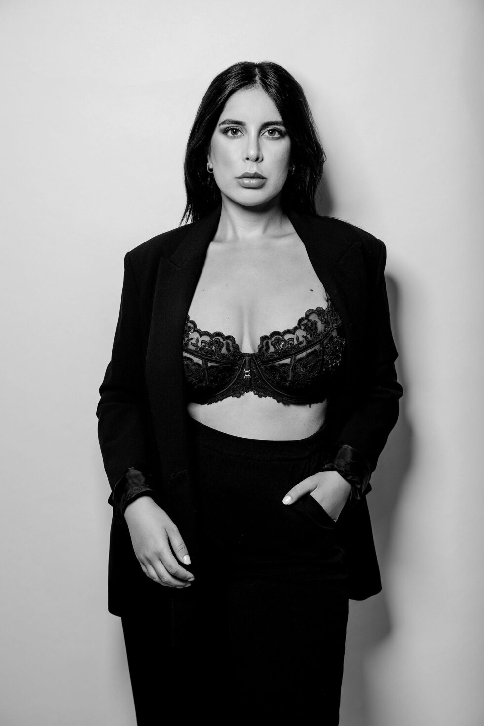 Schwarz-weiß Foto einer Frau in BH und Blazer, sie steht vor einer weißen Wand