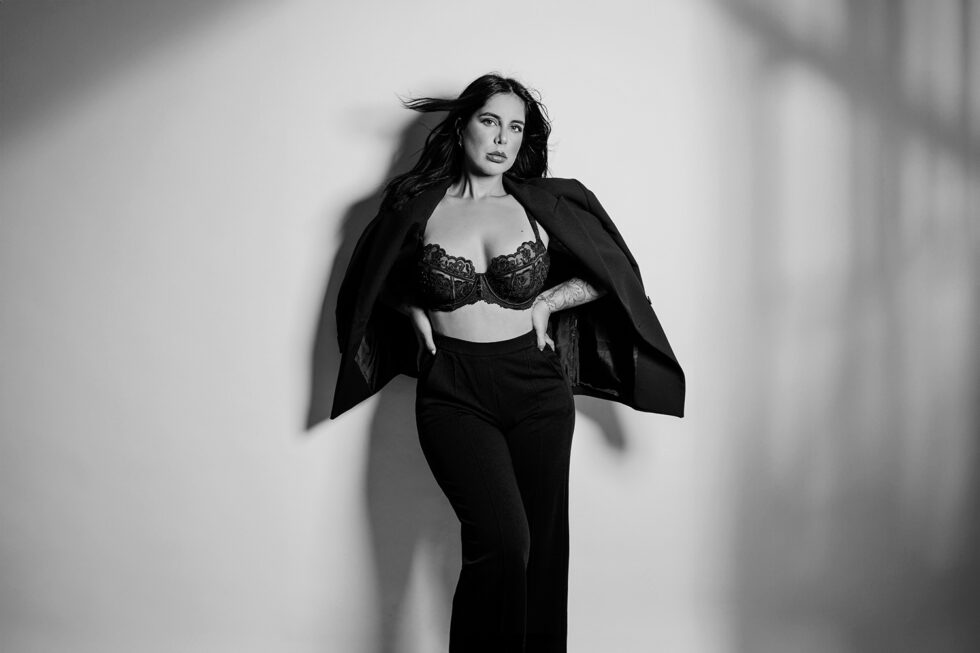 Schwarz-weiß Foto einer Frau in BH und Blazer, sie steht vor einer weißen Wand