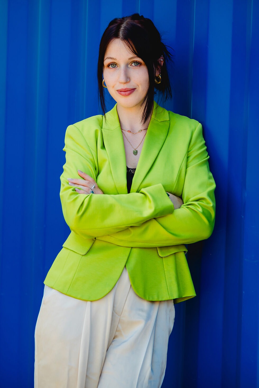 Junge Frau mit giftgrüne Blazer vor blauen Hintergrund steht mit verschränkten Armen da