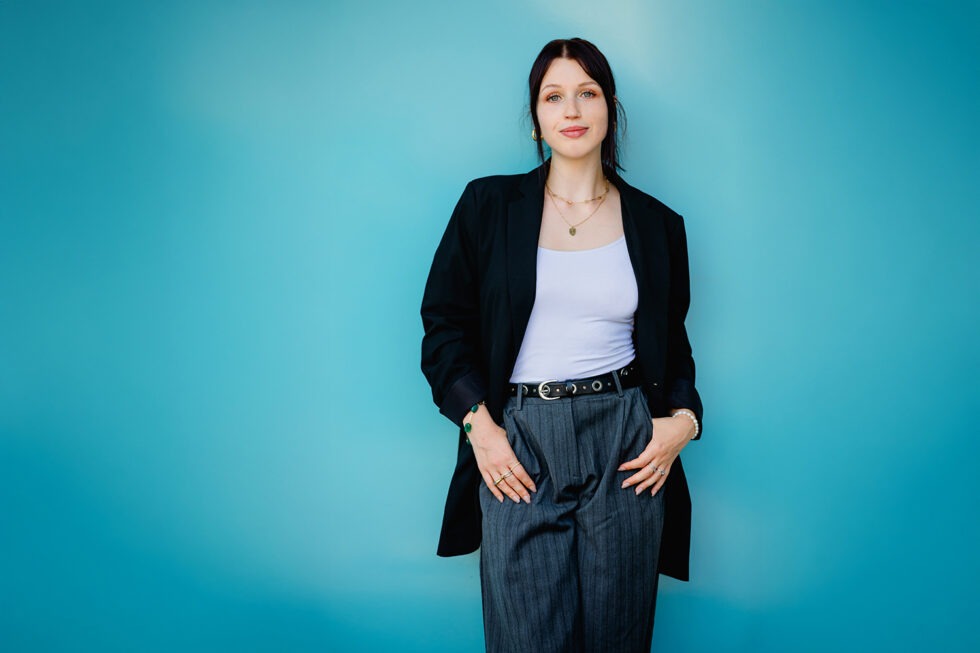 Portrait einer jungen Frau mit dunklen Haaren und Blazer, die vor einer blauen Wand steht