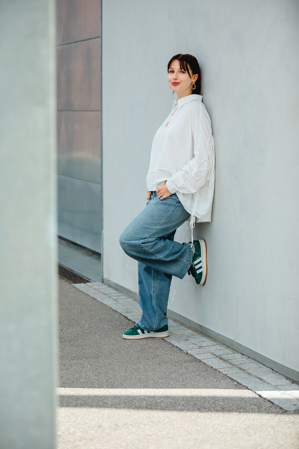 Eine junge Frau mit dunklen Haaren, lehnt an einer Betonwand und schaut in die Kamera