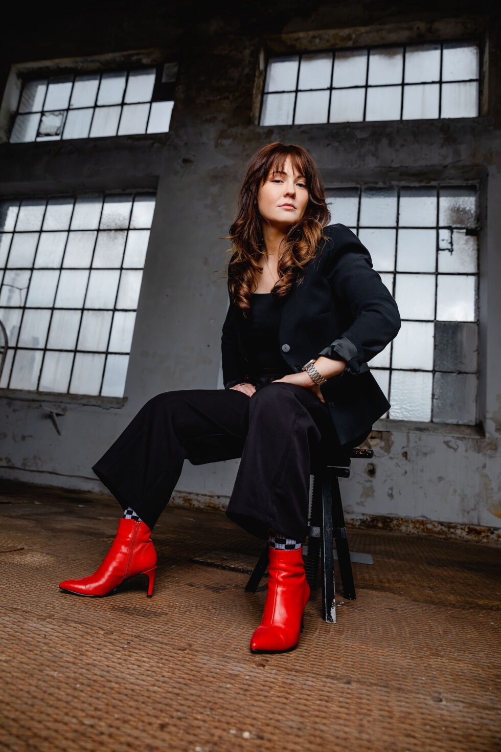 eine Frau im schwarzen Anzug mit roten Schuhen sitzt in einer Industriehallte